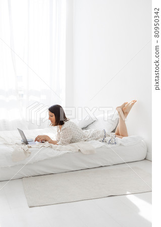 a young and beautiful European woman lying on a bed in a white bedroom with a laptop and working 80950342