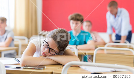 Teen girl with glasses sleeping at desk in classroom during lesson 80957195