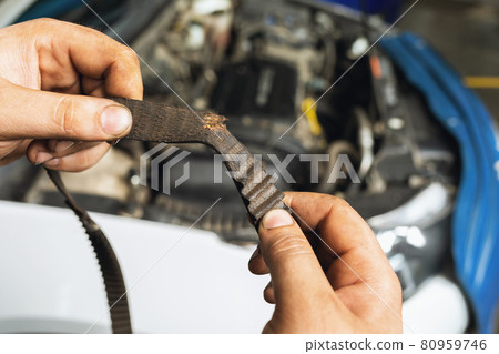 An auto mechanic shows a torn timing belt with worn teeth against the background of an open car hood close-up 80959746
