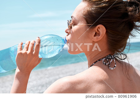 Young woman with sunglasses drinks water from plastic bottle on the beach. Hydration and healthy life in the summer. Wellness concept. Young woman with sunglasses drinks water from plastic bottle on the beach. Hydration and healthy life in the summer. Wellness concept. 80961906