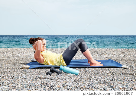 Slim Woman does exercises for abdominal muscles on the beach with blue sea in background. Strengthening and coordination of body. Practicing sport outdoors. Slim Woman does exercises for abdominal muscles on the beach with blue sea in background. Strengthening and coordination of body. Practicing sport outdoors. 80961907