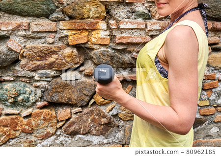 Slim Woman dressed in wet yellow sports tank top does execises with dumbblles outdoors with stone wall in background. Practicing sport. Wellbeing. Slim Woman dressed in wet yellow sports tank top does execises with dumbblles outdoors with stone wall in background. Practicing sport. Wellbeing. 80962185