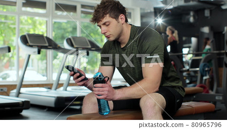 Portrait of confident bodybuilder looking phone screen at gym. Sportsman using phone and drinking water in gym. Man taking a break from his workout, drinking from water bottle. 80965796