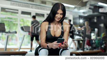 Beautiful athletic woman uses smartphone while resting on the bench after her Intensive fitness bodybuilding gym training. 80966000