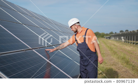 Male engineer in uniform walking and looking at solar power plant. Man in hard helmet examining object. Concept of solar station development and green energy. Worker on solar power station outdoor 80967119
