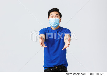 Different emotions, social distancing, self-quarantine on coronavirus and lifestyle concept. Happy asian man in medical mask and blue t-shirt, reaching hands towards camera to hold, catch something 80970425