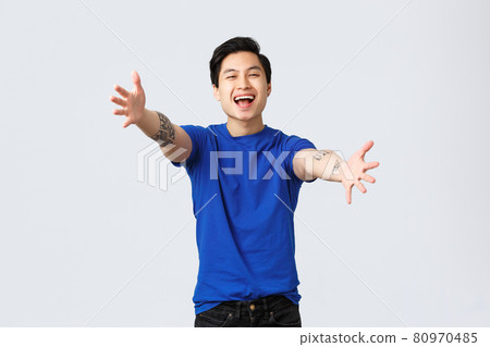 Cheerful friendly-looking young male student, asian man in blue t-shirt with tattoos, reaching hands towards camera for hug, want cuddles or embraces, greeting dear friend, grey background 80970485