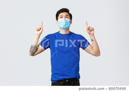 Different emotions, social distancing, self-quarantine and lifestyle concept. Disappointed and upset asian guy in medical mask and blue t-shirt, looking, pointing up with displeased grimace 80970498