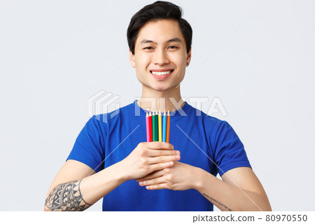 Self-quarantine, social distancine, leisure and lifestyle concept. Young handsome asian man with tattoo show colored pencils, found new hobby feeling happy, smiling at camera 80970550
