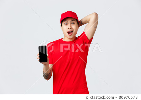 Nervous and troubled asian delivery guy in red cap and t-shirt uniform, gasping anxious, look confused and puzzled showing smartphone screen bad news, mixed-up order, stand grey background 80970578