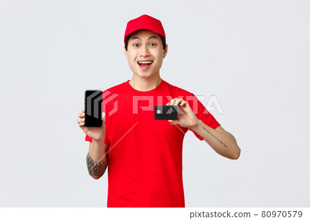 Delivery application, online orders and shipping concept. Quarantine wont stop from shopping. Cheerful asian courier in red cap and t-shirt, showing smartphone display and credit card 80970579