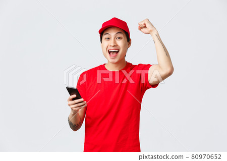 Happy, cheerful asian delivery guy in red uniform cap and t-shirt of carrier service, holding smartphone, reading good news, chanting with fist pump, yell yes celebrate victory or achivement 80970652
