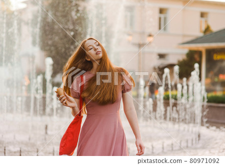 Young beautiful red-haired long-haired woman in a summer pink dress walks through the streets of a summer city. 80971092