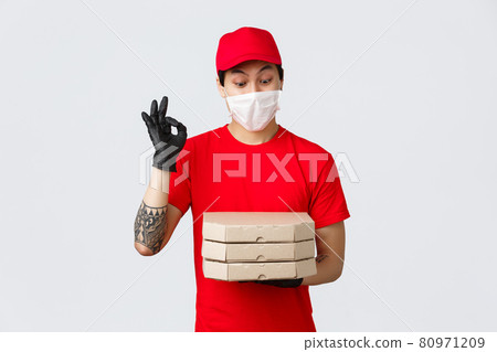 Amused asian delivery man red uniform, cap and t-shirt, wearing protective gloves and medical mask for client safety as deliver food to doorstep, bring pizza, show okay sign and stare at tasty pizza 80971209