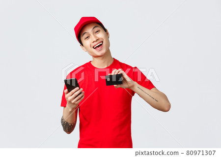 Friendly delivery guy in red uniform t-shirt and cap, showing smartphone screen and credit card, recommend application for tracking order online shopping. Courier advertise carrier service Friendly delivery guy in red uniform t-shirt and cap, showing smartphone screen and credit card, recommend application for tracking order online shopping. Courier advertise carrier service 80971307