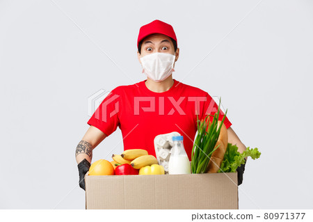 Online shopping, food delivery and coronavirus pandemic concept. Friendly asian delivery guy in medical mask and gloves, holding package with fresh groceries, courier bring products for customer 80971377