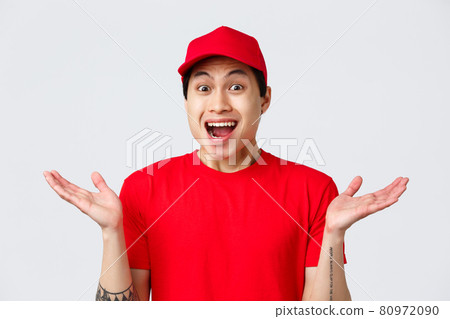 Delivery, contactless orders and shopping concept. Courier was surprised hearing great news. Delivery guy in red uniform cap and t-shirt, spread hands sideways and gasping amused, grey background 80972090