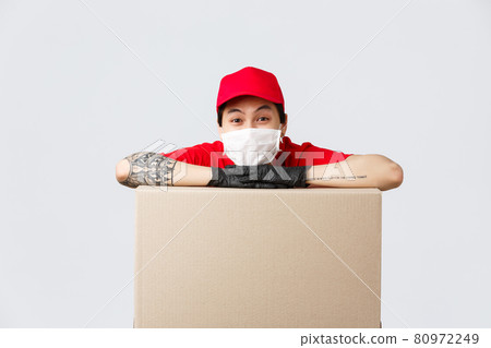 Delivery, online shopping and quarantine concept. Cheerful asian delivery guy in red cap uniform, medical mask and protective gloves, leaning on large box, order or parcel, promote courier service Delivery, online shopping and quarantine concept. Cheerful asian delivery guy in red cap uniform, medical mask and protective gloves, leaning on large box, order or parcel, promote courier service 80972249