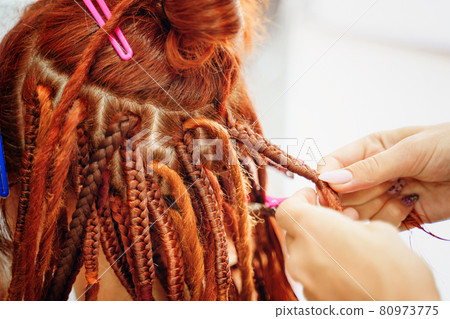 Hairdresser's hands braid girl's ginger dreadlocks. 80973775