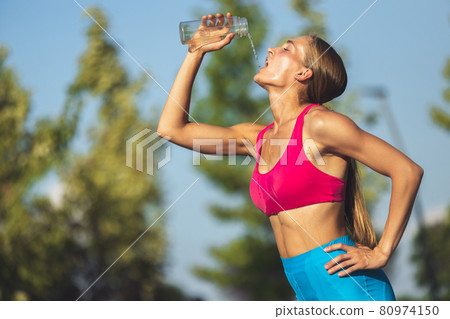 One young slim sportive caucasian woman in bright sportswear drinks water in the park or on city street. Side view 80974150