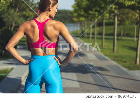 One young slim sportive caucasian woman in bright sportswear standing, looking aside in the park or on city street. 80974173