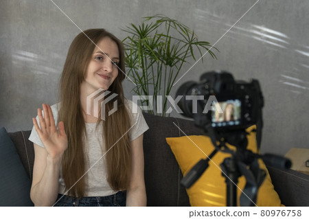 Young woman blogger takes video of herself on camera and waves her hand. Beautiful girl filming herself for vlogging Young woman blogger takes video of herself on camera and waves her hand. Beautiful girl filming herself for vlogging 80976758