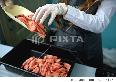 The fishmonger, vendor in fish department of a seafood market , stands behind the counter in a work apron and puts fresh frozen chilled red shrimp on a tray. Close-up The fishmonger, vendor in fish department of a seafood market , stands behind the counter in a work apron and puts fresh frozen chilled red shrimp on a tray. Close-up 80987147