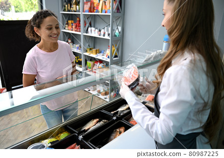 Smiling African American woman, customer at seafood store buying fish. Fishmonger serves fresh red salmon fillet steak Smiling African American woman, customer at seafood store buying fish. Fishmonger serves fresh red salmon fillet steak 80987225