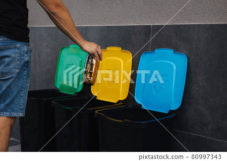 Close-up of human hand throwing empty plastic water bottle in recycling bin. Tree recycling bins outdoors 80997343