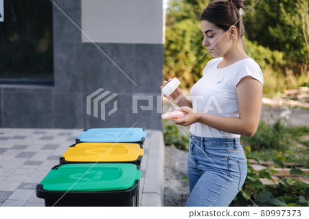 Woman throwing out in recycling bin washed and empty shampoo and deodorant bottles. Female looks at the packages to find out in wich recycling bin throw the garbage 80997373