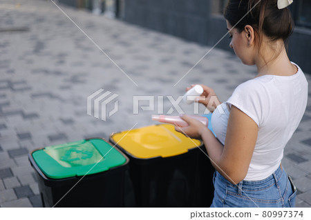 Woman throwing out in recycling bin washed and empty shampoo and deodorant bottles. Female looks at the packages to find out in wich recycling bin throw the garbage 80997374