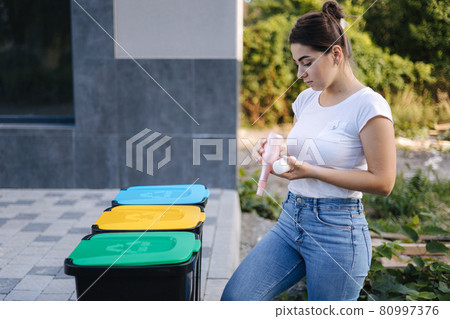 Woman throwing out in recycling bin washed and empty shampoo and deodorant bottles. Female looks at the packages to find out in wich recycling bin throw the garbage 80997376