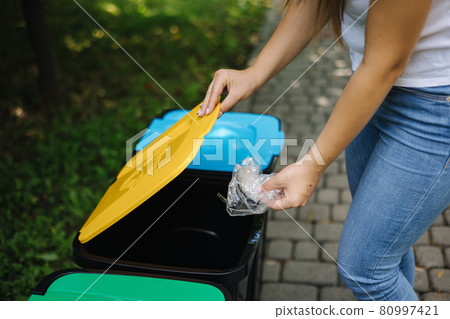 Female in the park throwing plastic bag into recycling bin. Different colours on plastic bins. Green, blue and yellow Female in the park throwing plastic bag into recycling bin. Different colours on plastic bins. Green, blue and yellow 80997421