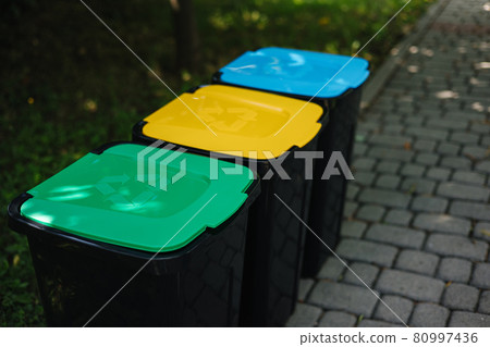 Outdoors in the park is recycling bins different colours. Tree closed plastic bins. Green, yeallow and blue Outdoors in the park is recycling bins different colours. Tree closed plastic bins. Green, yeallow and blue 80997436