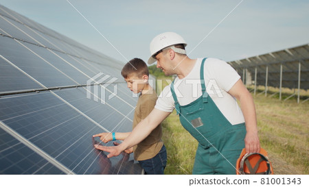 An young engineer father is explaining to his little son an operation and performance of photovoltaic solar panels at sunset. Concept: renewable energy, technology, electricity, green, future, family. An young engineer father is explaining to his little son an operation and performance of photovoltaic solar panels at sunset. Concept: renewable energy, technology, electricity, green, future, family. 81001343