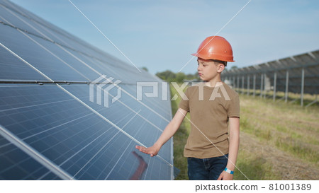 Portrait of a child near the solar panels. A little boy in a protective helmet touches the solar panels with his hand. Shooting at a solar power plant. Ecological farm. Solar power station. People Portrait of a child near the solar panels. A little boy in a protective helmet touches the solar panels with his hand. Shooting at a solar power plant. Ecological farm. Solar power station. People 81001389