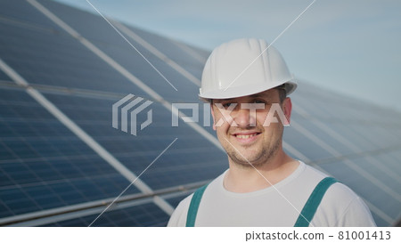 Portrait of happy male engineer in protective helmet looking to camera. Handsome man in uniform smiling while standing at solar power farm. Concept of green energy. Ecological future. Portrait of happy male engineer in protective helmet looking to camera. Handsome man in uniform smiling while standing at solar power farm. Concept of green energy. Ecological future. 81001413