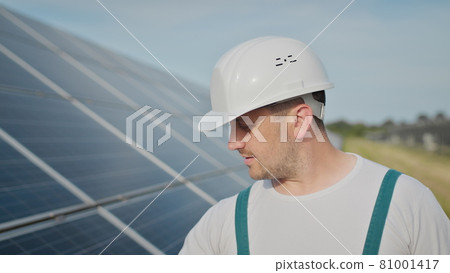 Portrait of happy male engineer in protective helmet looking to camera. Handsome man in uniform smiling while standing at solar power farm. Concept of green energy. Ecological future. 81001417