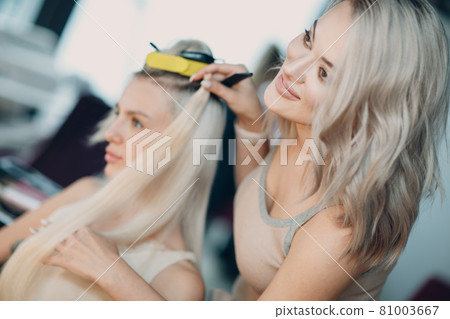 Hairdresser female making hair extensions to young woman with blonde hair in beauty salon. Professional hair extension. Hairdresser female making hair extensions to young woman with blonde hair in beauty salon. Professional hair extension. 81003667