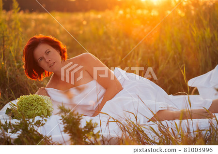 Charming woman in white bed in a field at sunset in summer looking at the camera 81003996