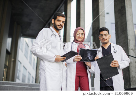 Malay doctors wearing white coats and scrubs, posing together, standing outside the hospital Malay doctors wearing white coats and scrubs, posing together, standing outside the hospital 81009969