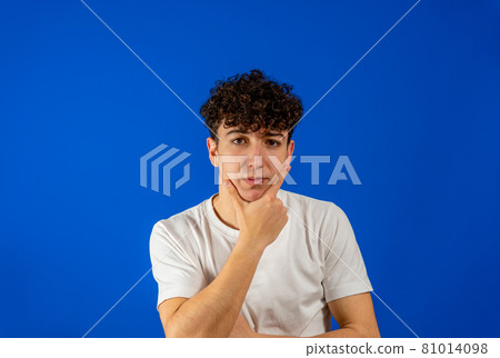 Young caucasian man with curly hair with his hand on his chin , he is pensive and doubts in the face of the adversities that life will present her. Isolated on blue studio background. Doubt concept Young caucasian man with curly hair with his hand on his chin , he is pensive and doubts in the face of the adversities that life will present her. Isolated on blue studio background. Doubt concept 81014098