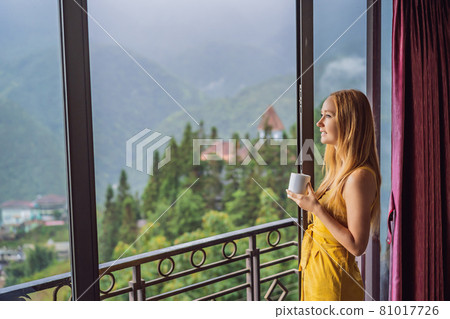 A young woman holding coffee cup while sittinhg on chair on balcony , looking at mountains and green nature. Woman in Sapa in the fog, Northwest Vietnam. Vietnam travel concept. UNESCO heritage 81017726