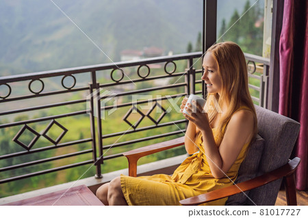 A young woman holding coffee cup while sittinhg on chair on balcony , looking at mountains and green nature. Woman in Sapa in the fog, Northwest Vietnam. Vietnam travel concept. UNESCO heritage 81017727