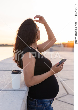 Pregnant hispanic woman holds smartphone and relax on embankment with warm light in morning Pregnant hispanic woman holds smartphone and relax on embankment with warm light in morning 81023393