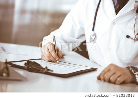 Unknown male doctor sitting and working with clipboard of medication history record in clinic at his working place, close-up. Young physician at work. Perfect medical service, medicine concept Unknown male doctor sitting and working with clipboard of medication history record in clinic at his working place, close-up. Young physician at work. Perfect medical service, medicine concept 81028818