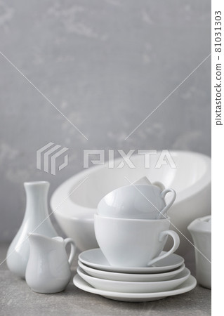 Empty crockery set or ceramic dishes. White kitchen dishware and tableware on table near grey wall Empty crockery set or ceramic dishes. White kitchen dishware and tableware on table near grey wall 81031303