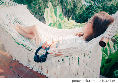 Beautiful girl in a white hammock in the garden at home. Young woman listening to music & dreaming of summer nature. Concept of freedom of happiness on vacation and relaxing. hand holding headphones. 81032146