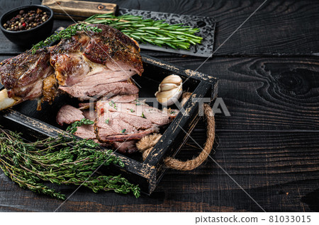 Roasted mutton lamb leg sliced in a wooden tray with meat cleaver. Black wooden background. Top view. Copy space 81033015