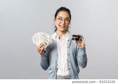 Business, finance and investment concept. Thoughtful smiling young asian rich woman in glasses, bite lip curious, look up while thinking what buy on her prize, hold money dollars and credit card Business, finance and investment concept. Thoughtful smiling young asian rich woman in glasses, bite lip curious, look up while thinking what buy on her prize, hold money dollars and credit card 81033226